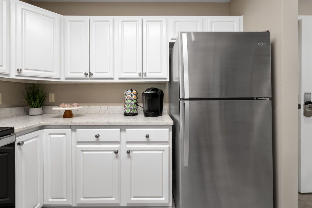 A modern kitchen in Hilliard, Ohio features white cabinets, a stainless steel refrigerator, a coffee maker with a pod holder, a plant, and a cake stand on the marble countertop—perfect for comfortable senior living.