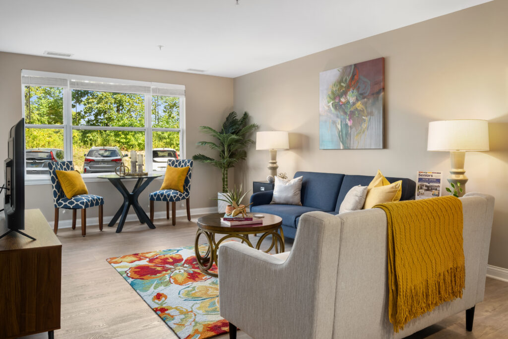 A bright, cozy living room in Hilliard, Ohio features a blue sofa, beige armchair with a yellow blanket, colorful rug, round coffee table, two patterned chairs at a small table by a large window—perfect for luxury independent living.