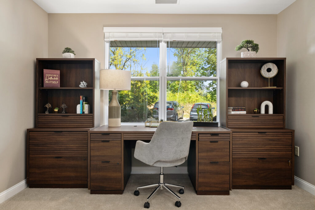 A modern home office in Hilliard, Ohio, features a wooden desk and matching shelves, a gray swivel chair, a desk lamp, and decor—perfect for independent living. A large window behind the desk lets in natural light and shows greenery outside.