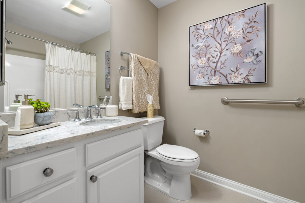 A clean bathroom with white cabinets, a marble countertop, and floral artwork above the toilet offers luxury and comfort—perfect for independent living. Beige walls, a towel rack, grab bar, and a shower curtain complete this inviting space.