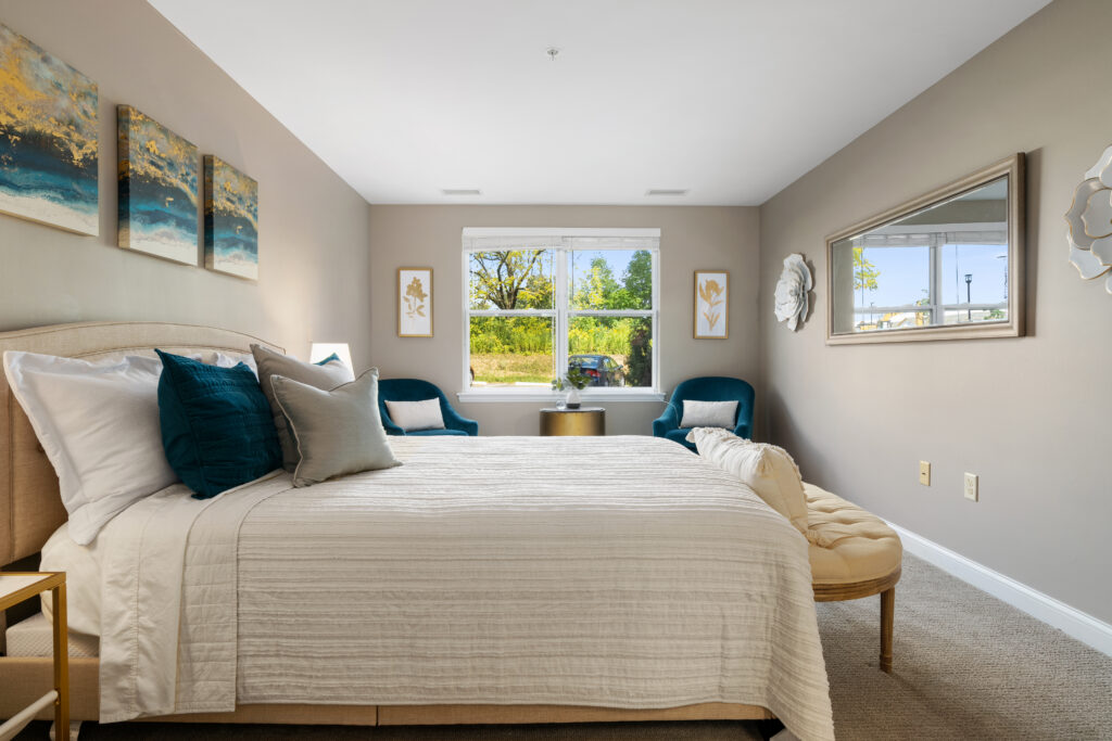 A bright bedroom designed for luxury independent living, featuring a large bed with white and teal bedding, two blue chairs by a window, a round table, wall art, a large mirror, and neutral-colored walls and carpet.