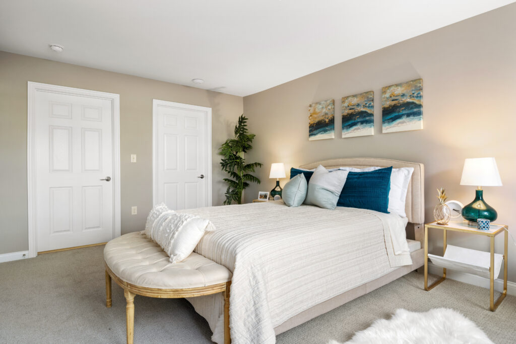 Bright, modern bedroom designed for luxury senior living, featuring a beige bed with blue accent pillows, a bench at the foot, stylish nightstands with lamps, wall art above the headboard, two doors, and a potted plant in the background.