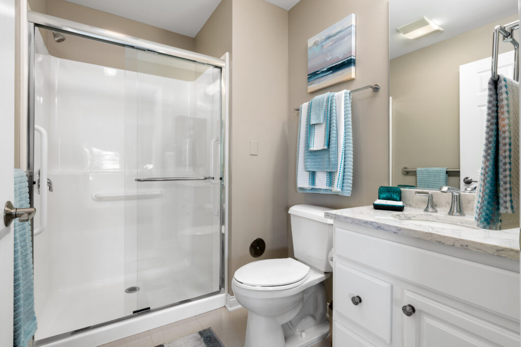 Modern bathroom with glass shower, white toilet, marble countertop, and white vanity. Located in Hilliard Ohio senior living, it features light tan walls, blue and white towels, wall art, and a mirror above the sink—bright and luxury feel.
