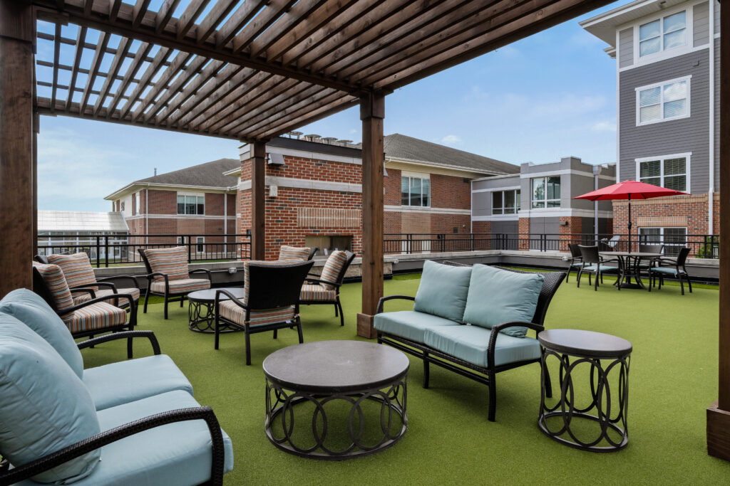 Luxury outdoor patio area with cushioned chairs, sofas, and round tables under a wooden pergola on green artificial turf. Enjoy independent living in Hilliard Ohio, surrounded by apartment buildings and vibrant seating with striped chairs and a red umbrella.