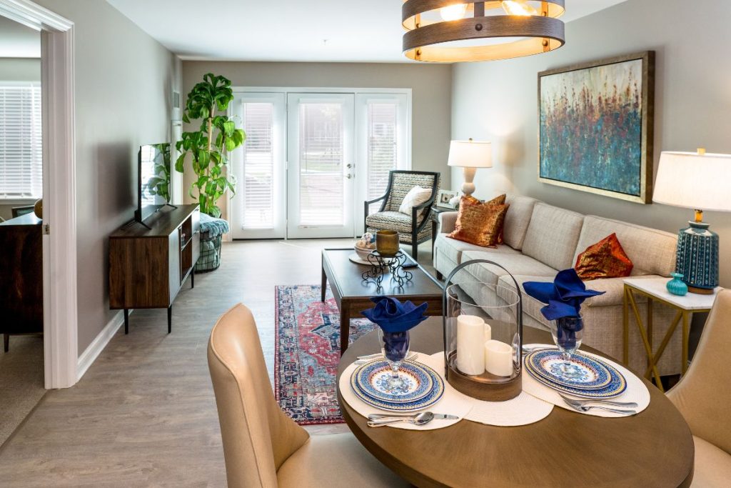 A modern, cozy living-dining room in Hilliard, Ohio features a round table set for two, a beige sofa with colorful pillows, a large plant, artwork on the wall, and French doors letting in natural light—a touch of luxury for independent living.