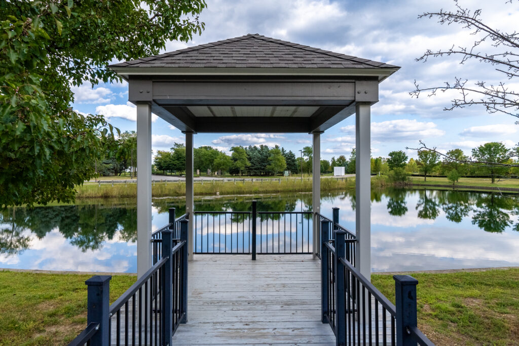 A wooden gazebo with a ramp overlooks a calm pond surrounded by grass and trees in Hilliard, Ohio, offering a peaceful retreat for senior living under a blue sky with clouds reflected in the water.