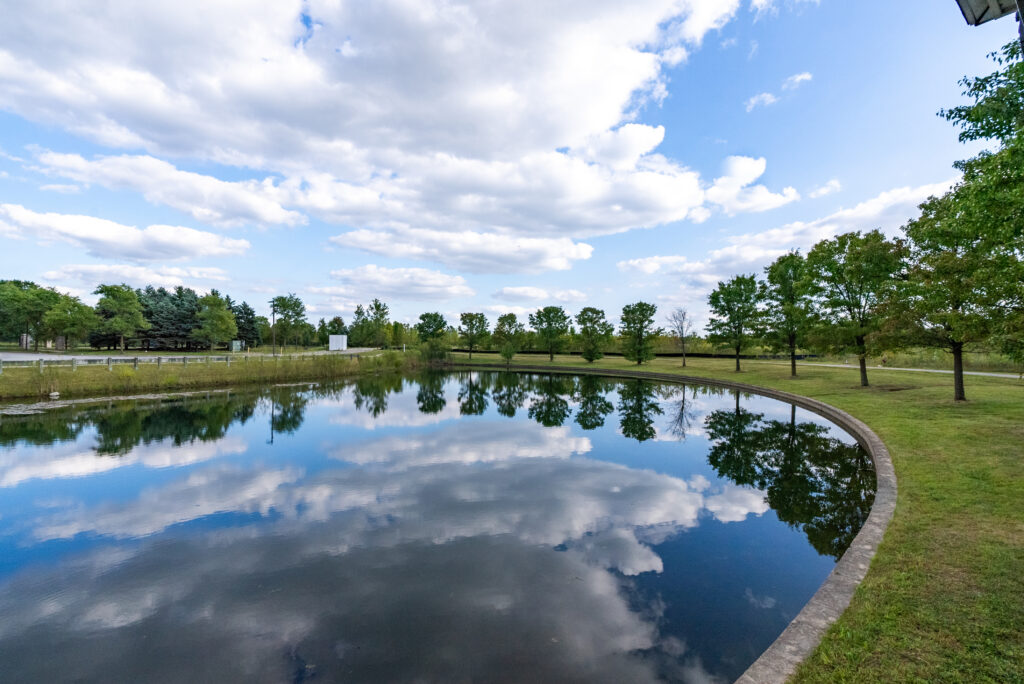 A curved pond reflects a blue sky with scattered clouds, surrounded by grassy areas and trees lining the shore—creating a serene and peaceful outdoor scene ideal for senior living.