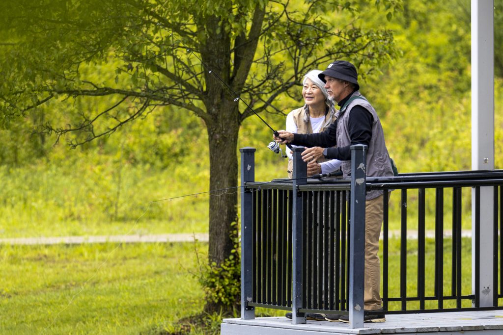 An older couple stands on a small platform by a black railing, fishing together in a lush green park—a perfect day for senior living in Hilliard, Ohio. The man holds a fishing rod while the woman smiles beside him in casual outdoor clothing.