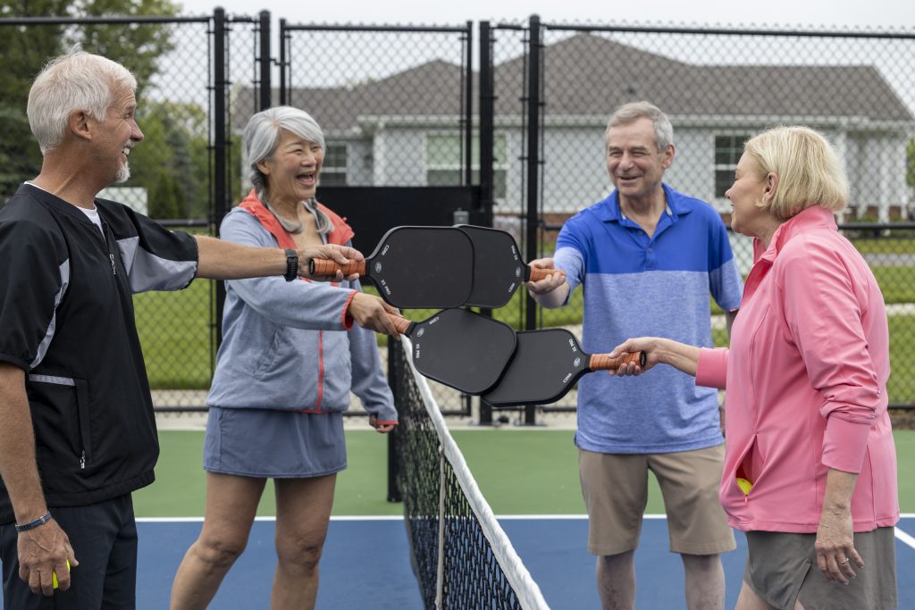 Four seniors, two men and two women, stand on an outdoor pickleball court at a senior living community, smiling and tapping their paddles together over the net in a friendly gesture before a game.
