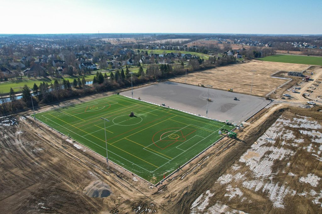 Aerial view of a newly constructed sports field with artificial turf, set near a luxury independent living community, surrounded by dirt and construction areas, adjacent to residential neighborhoods and open fields under a clear sky.