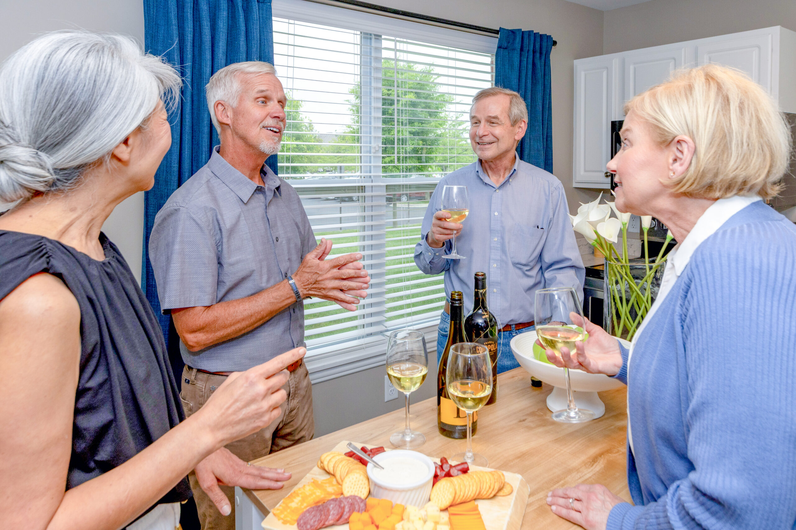 Four older adults stand around a kitchen island in a luxury independent living setting, chatting and smiling with glasses of white wine. A platter of cheese, crackers, and meat is on the counter, with wine bottles and glasses in the foreground.