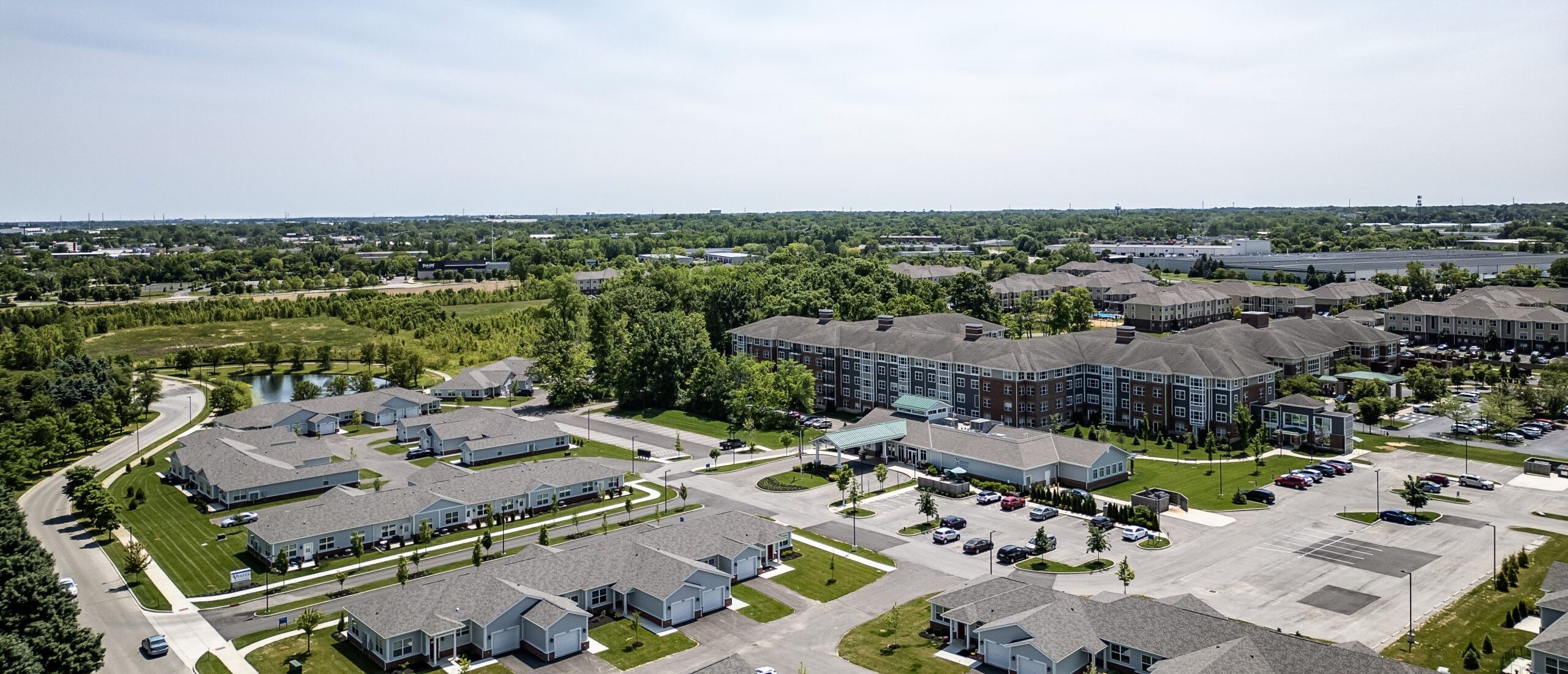 Aerial view of a suburban neighborhood with rows of houses, a senior living apartment building, green lawns, trees, roads, and parked cars on a bright day. Open fields and distant buildings are visible in the background.