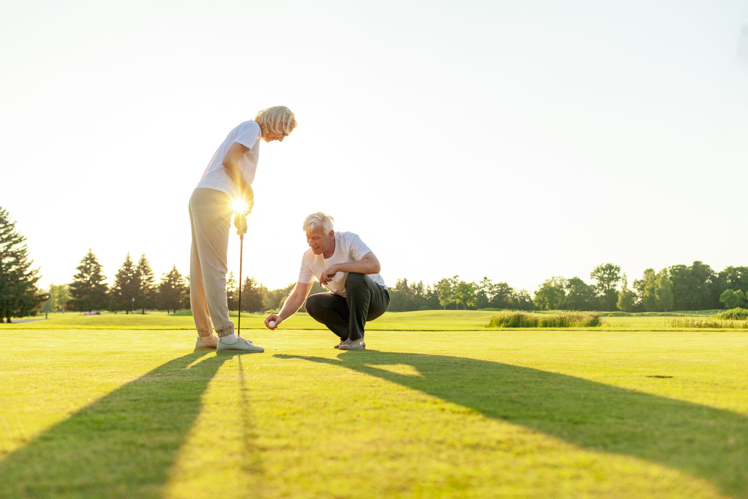 Two seniors enjoy a sunny day on a luxury golf course; one stands with a club while the other squats to place a golf ball, both smiling. Long shadows stretch across the lush green grass, reflecting vibrant senior living.