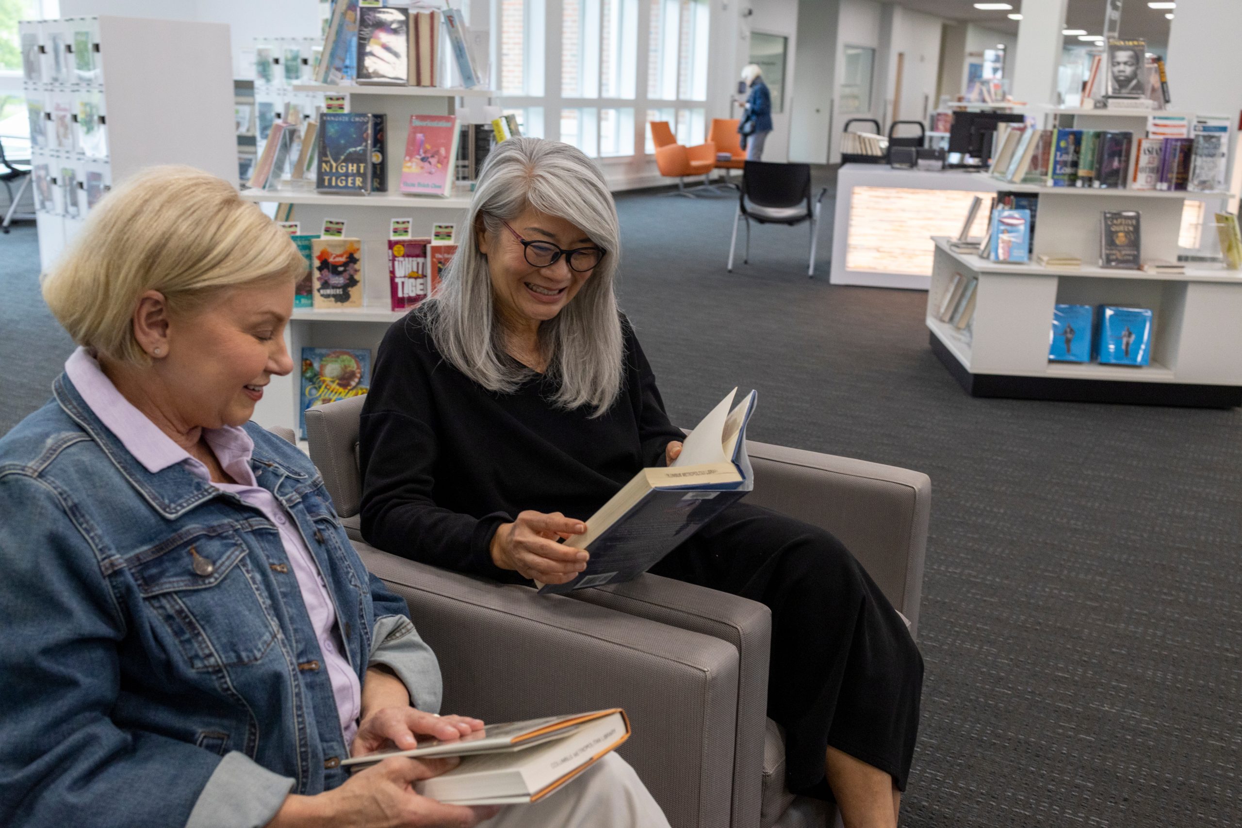 Two senior women sit in armchairs at a library in Hilliard, Ohio, smiling and reading books. Shelves filled with books and magazines are visible in the background, creating a bright and welcoming atmosphere.