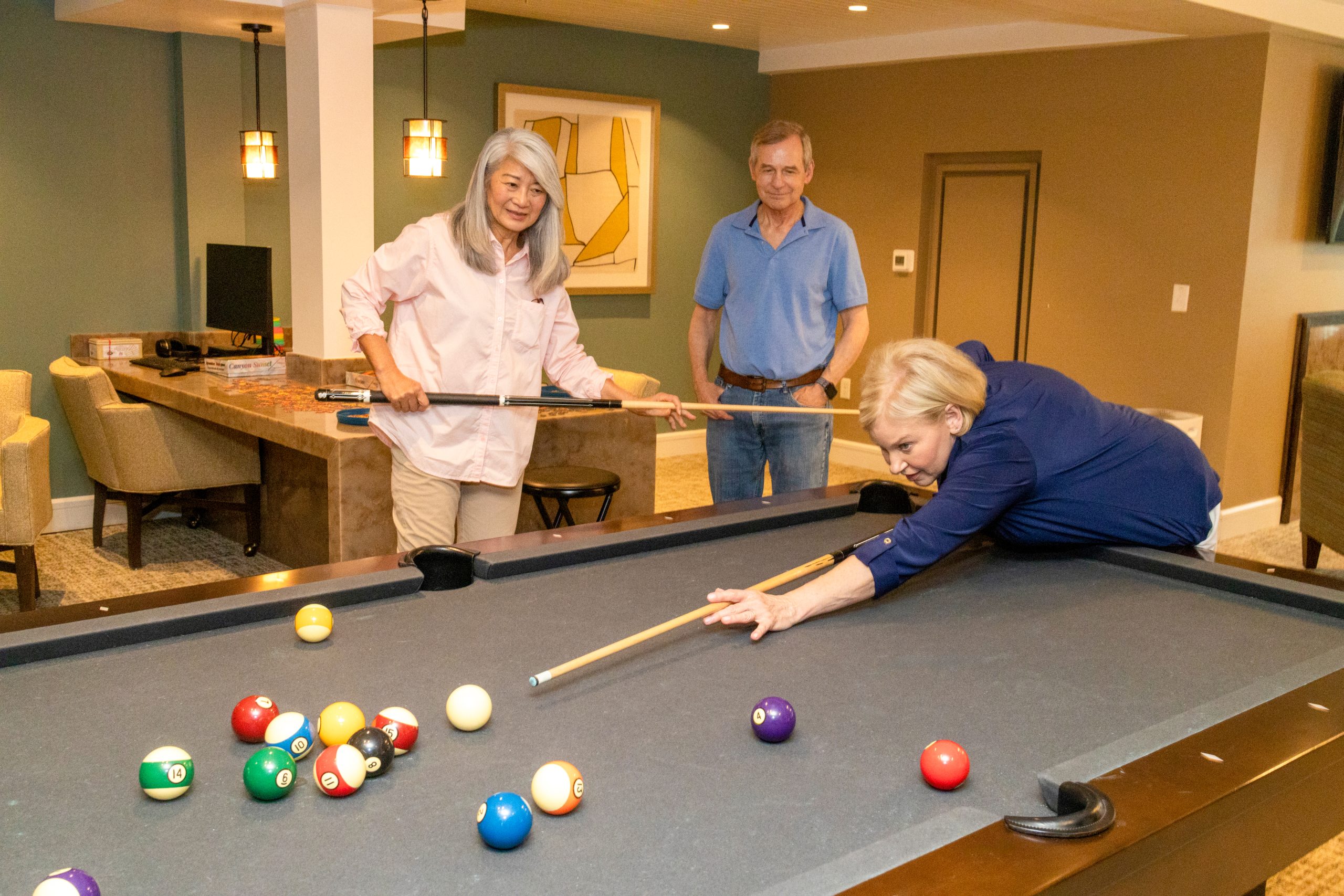 Three people enjoy a game of pool in a warmly lit room. In this luxury senior living space, a woman takes her shot while another stands with a cue stick, and a man in blue watches and smiles.