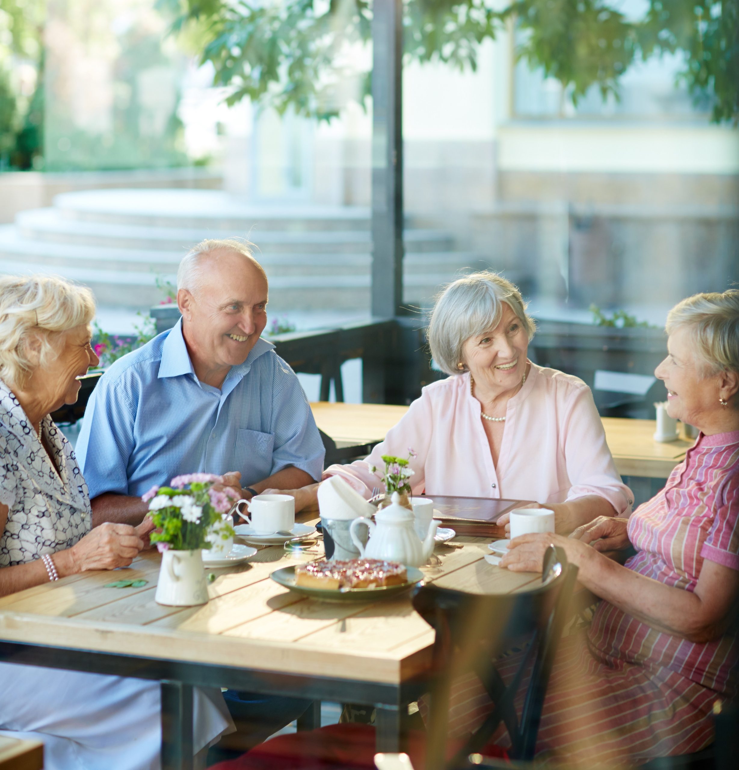 Four smiling older adults sit around a table at an outdoor café in Hilliard, Ohio, enjoying tea and pastries together. The table is decorated with small flowers, enhancing the cheerful atmosphere of this luxury senior living experience.