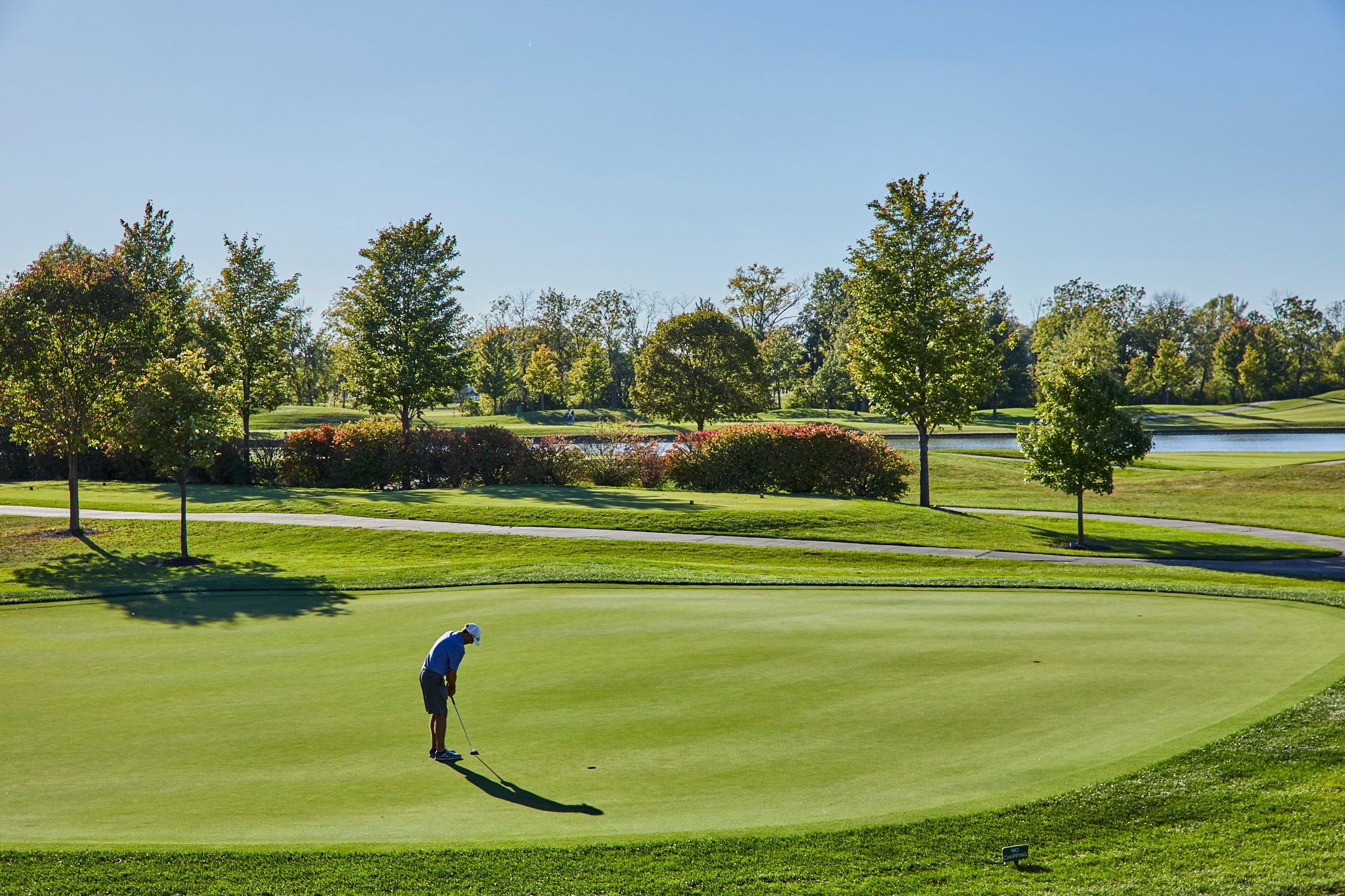 A golfer in a blue shirt and white hat prepares to putt on a sunny, green golf course surrounded by trees and a pond—the perfect setting for luxury independent living.