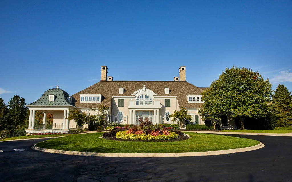 A large, elegant house in Hilliard, Ohio with a symmetrical facade, two chimneys, and a manicured lawn featuring colorful flowerbeds—perfect for senior independent living. Trees and a gazebo flank the building under a clear blue sky.
