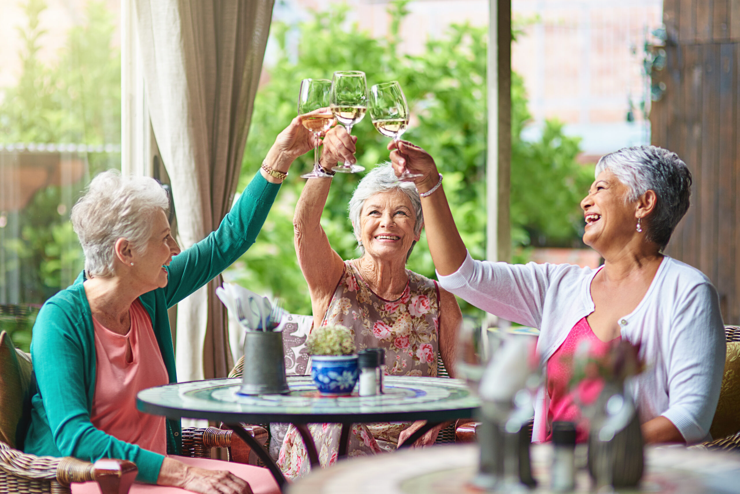 Three senior women sit at an outdoor table, smiling and raising glasses of wine in a toast. The bright, cheerful setting with lush greenery reflects the joy and connection found in independent living.