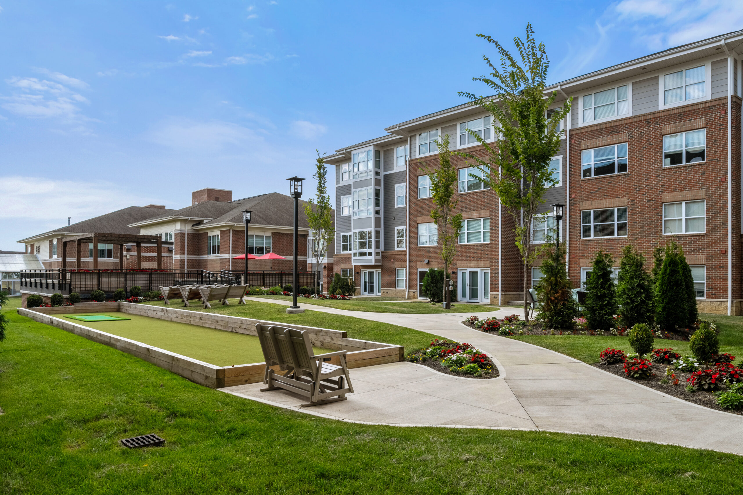 A modern brick apartment building for independent living with large windows, landscaped gardens, a walking path, benches, and a bocce ball court—perfect for luxury senior living on a bright, sunny day.