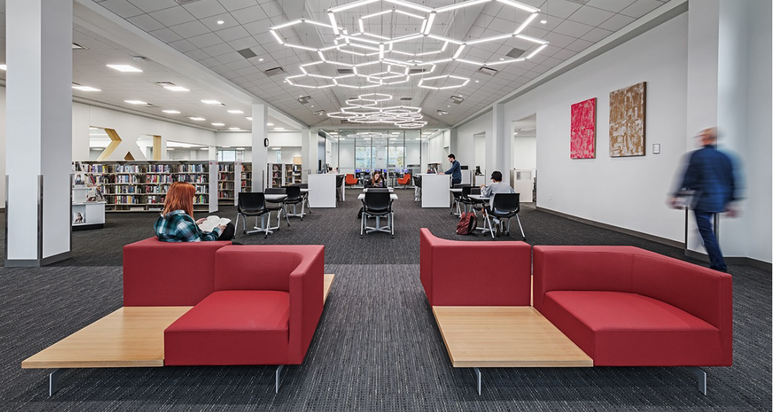 A modern library interior in Hilliard Ohio with red sofas, bookshelves, tables, and seniors reading or walking. Geometric ceiling lights and colorful wall art enhance the bright, open atmosphere for a touch of luxury.