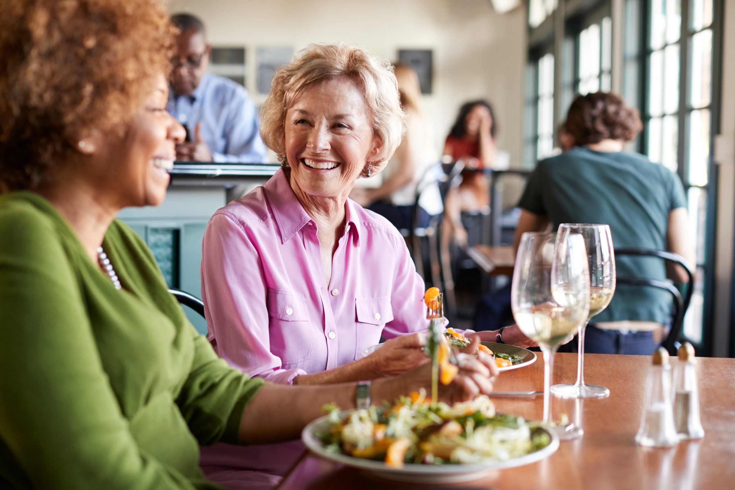 Two women enjoy a lively meal at a stylish restaurant in Hilliard, Ohio, smiling over salads and white wine. The friendly atmosphere hints at the vibrant social life found in luxury senior living communities.