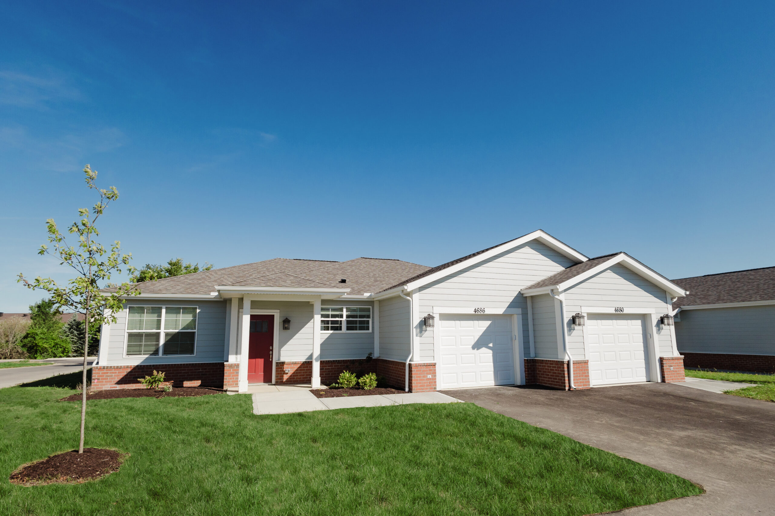 A single-story suburban house in Hilliard, Ohio with white siding, a red front door, two white garage doors, a gray roof, and a well-maintained lawn with a young tree offers luxury independent living under a clear blue sky.