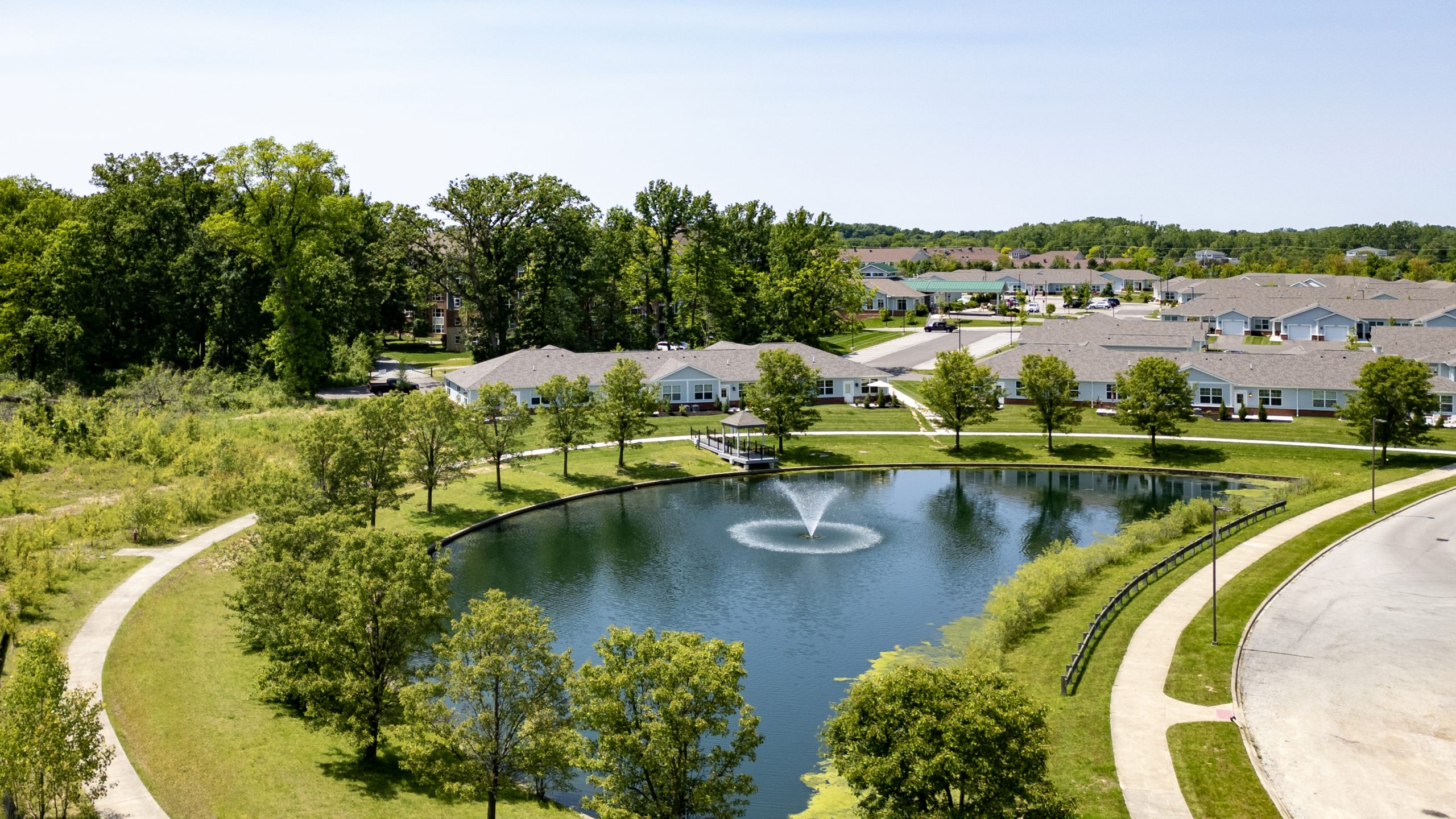 A small pond with a central fountain is surrounded by walking paths, trees, and luxury single-story residential buildings under a clear sky.