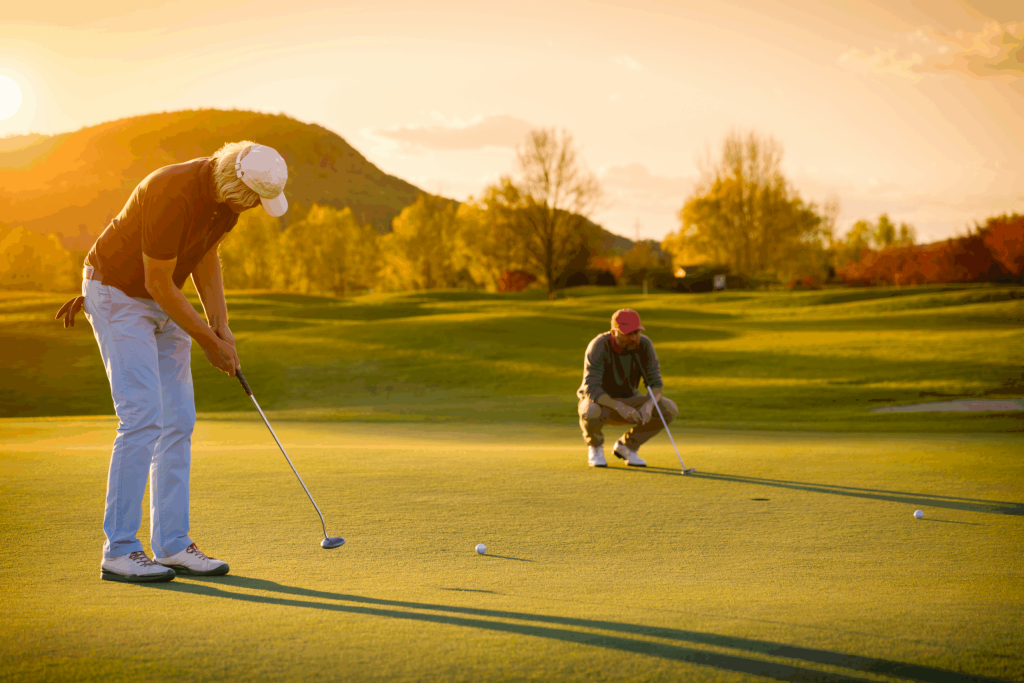 Two seniors enjoying a round of golf at sunset on a green field in Hilliard, Ohio; one golfer is putting while the other crouches behind, observing, with long shadows and trees in the background—celebrating independent living.