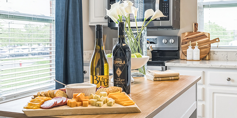 A wooden kitchen island in this luxury space holds a cheese and cracker platter, wine, and green apples. White calla lilies brighten the senior living kitchen, with white cabinets and a window offering charming views outside.