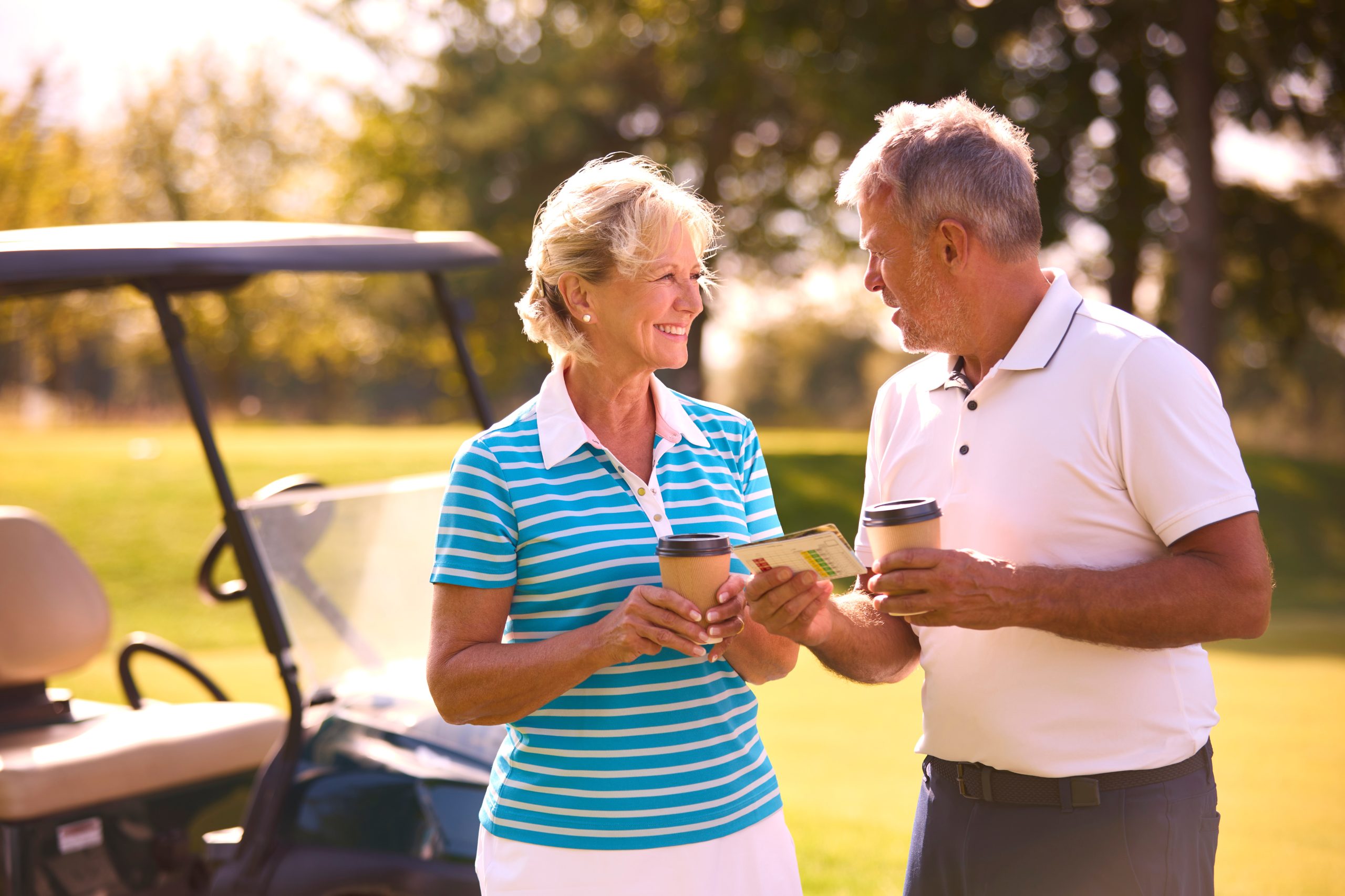 A smiling senior couple stands next to a golf cart on a sunny golf course in Hilliard, Ohio, chatting and holding drinks. The woman holds a scorecard, showing how independent living can be filled with fun and active moments surrounded by greenery.