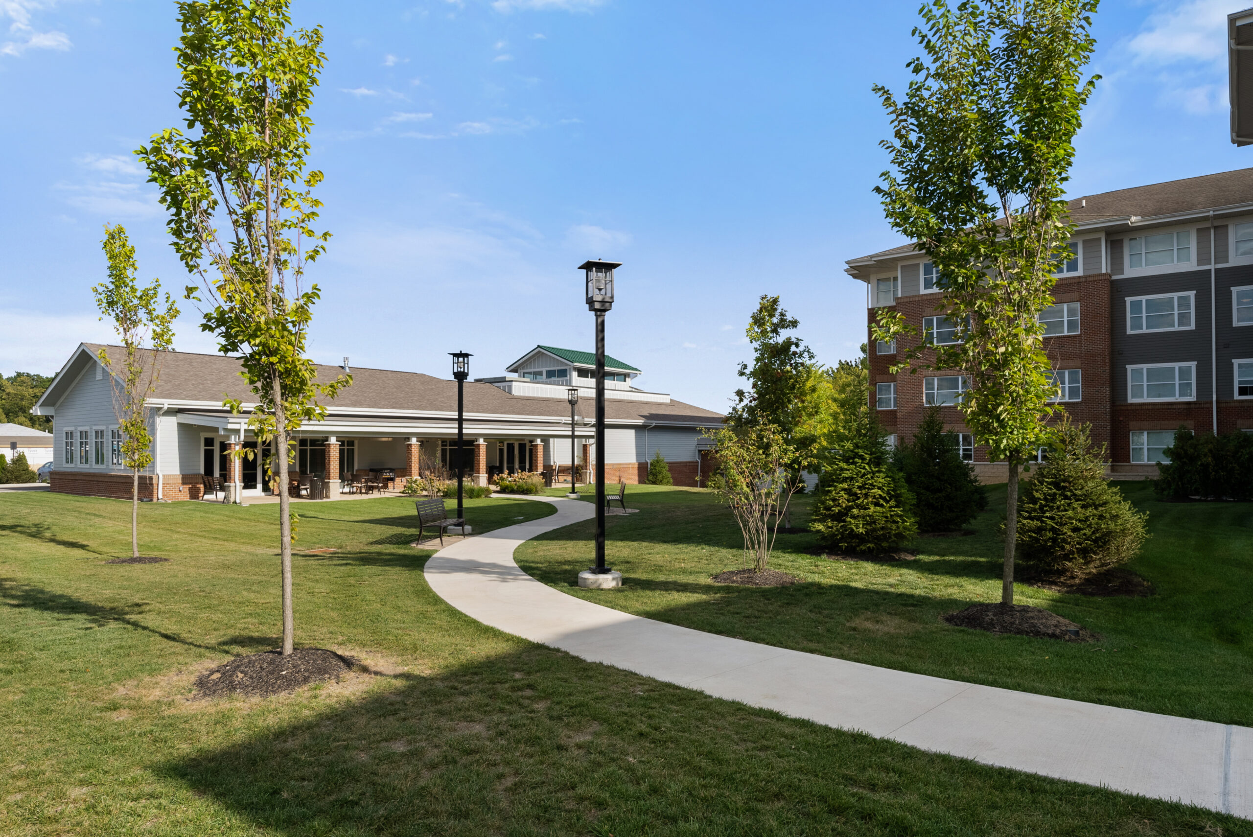 A curved sidewalk winds through a green lawn with young trees and street lamps, leading to luxury buildings with brick and siding exteriors under a clear blue sky in Hilliard’s independent living community.