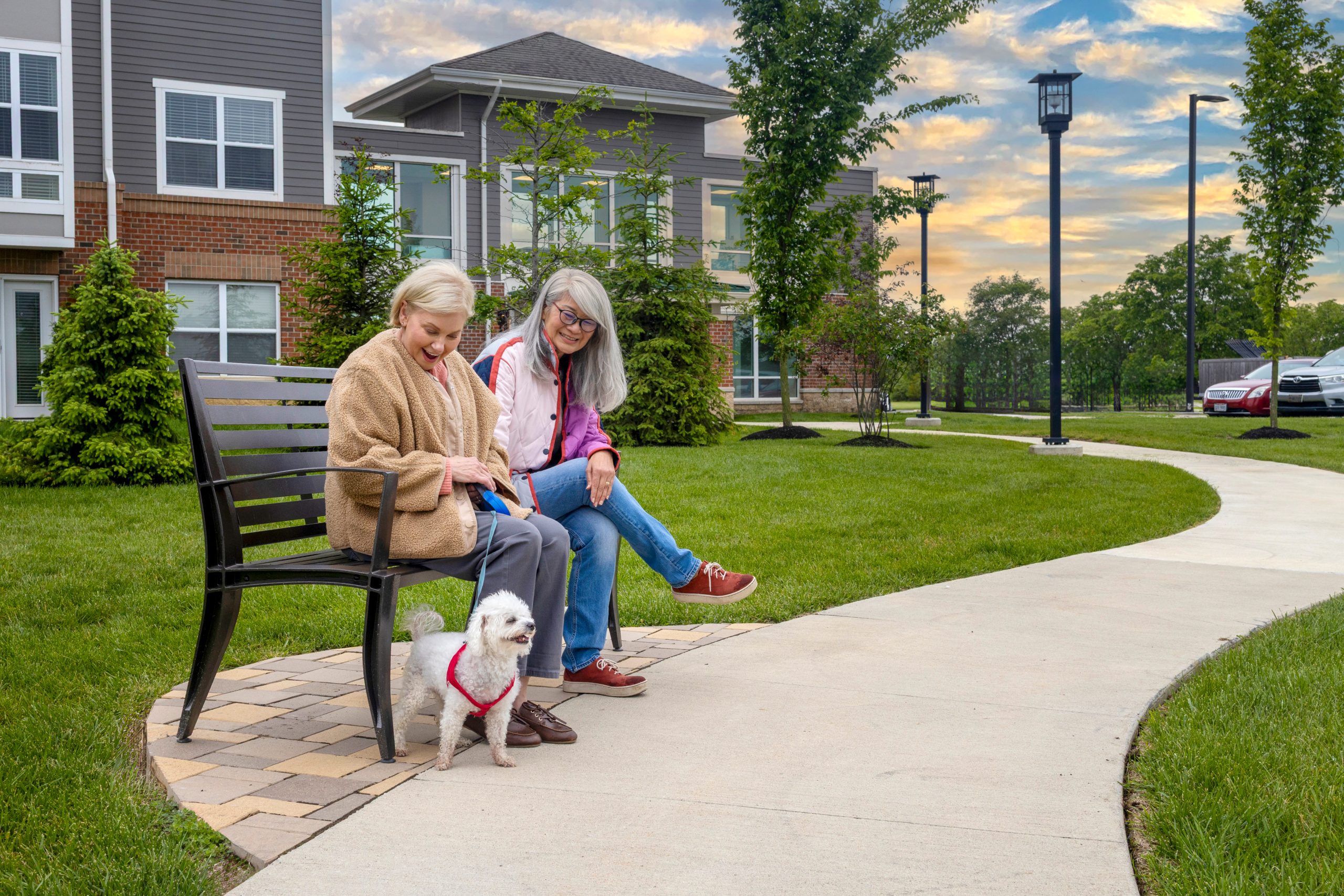 Two older women enjoy senior living on a bench in Hilliard, Ohio, smiling at a small white dog in a red harness. They relax by an apartment building as the sun sets and clouds drift across the sky, embracing moments of luxury together.