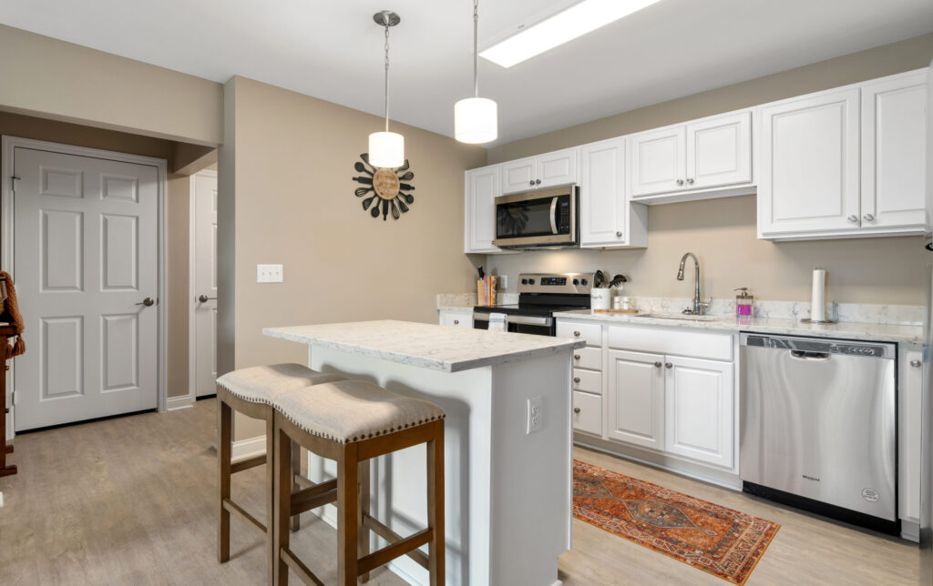 Modern kitchen with white cabinets, stainless steel appliances, an island with two stools, pendant lights, a colorful rug, and neutral walls. There is a closed white door and wooden stairs visible on the left.