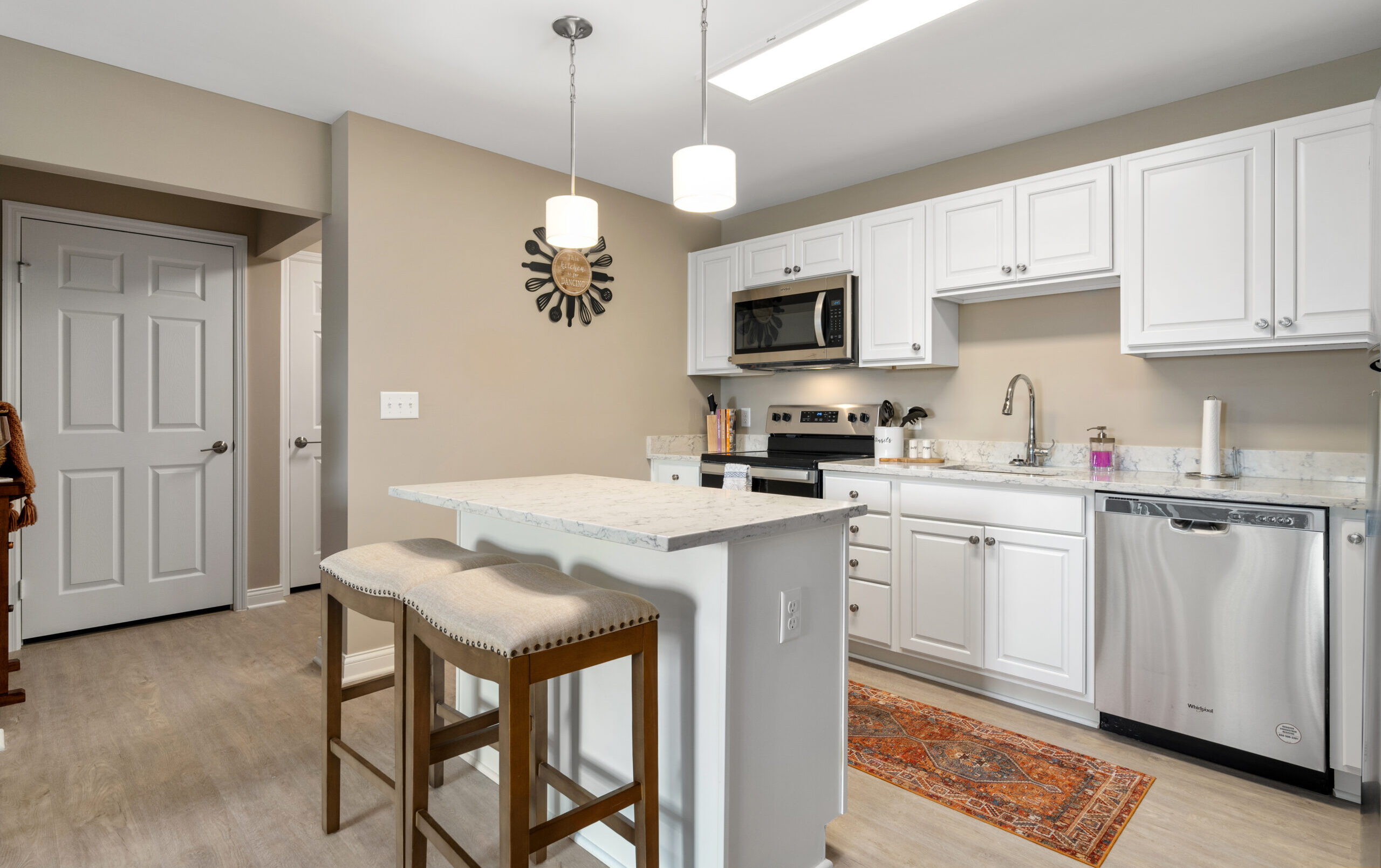 Modern kitchen with white cabinets, stainless steel appliances, an island with two stools, pendant lights, a colorful rug, and neutral walls. There is a closed white door and wooden stairs visible on the left.