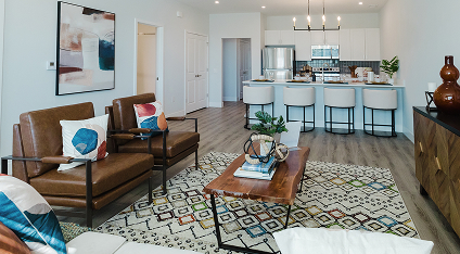 Modern living room in luxury apartments with brown leather chairs, a patterned rug, a wooden coffee table, and a kitchen featuring white barstools and stainless steel appliances in the background.