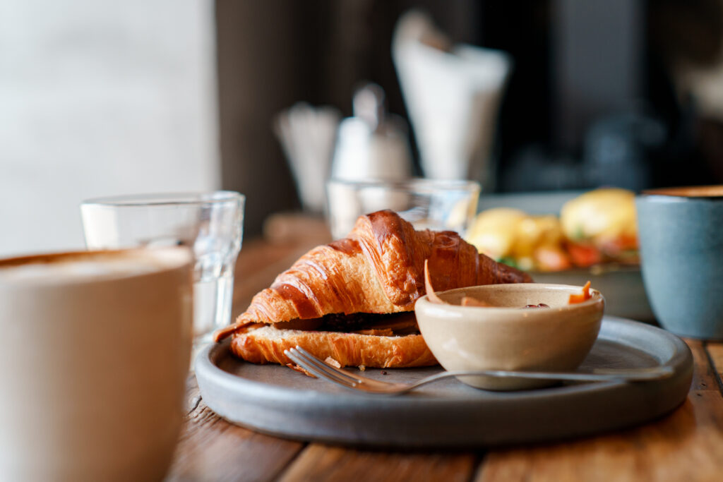 A croissant on a gray plate with a small bowl, fork, and cup of coffee on a wooden table. Glasses of water and blurred breakfast foods are visible in the background.