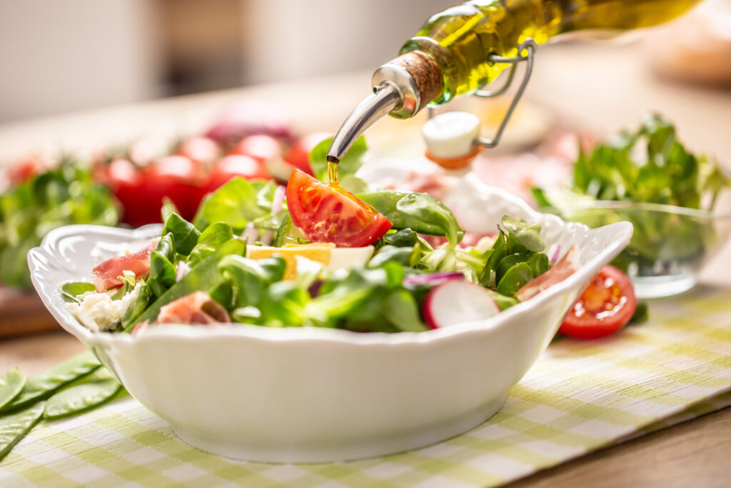 A close-up of a fresh salad with spinach, tomatoes, radishes, and cheese in a white bowl, as olive oil is being poured on top. The bowl sits on a checkered cloth, with vegetables blurred in the background.