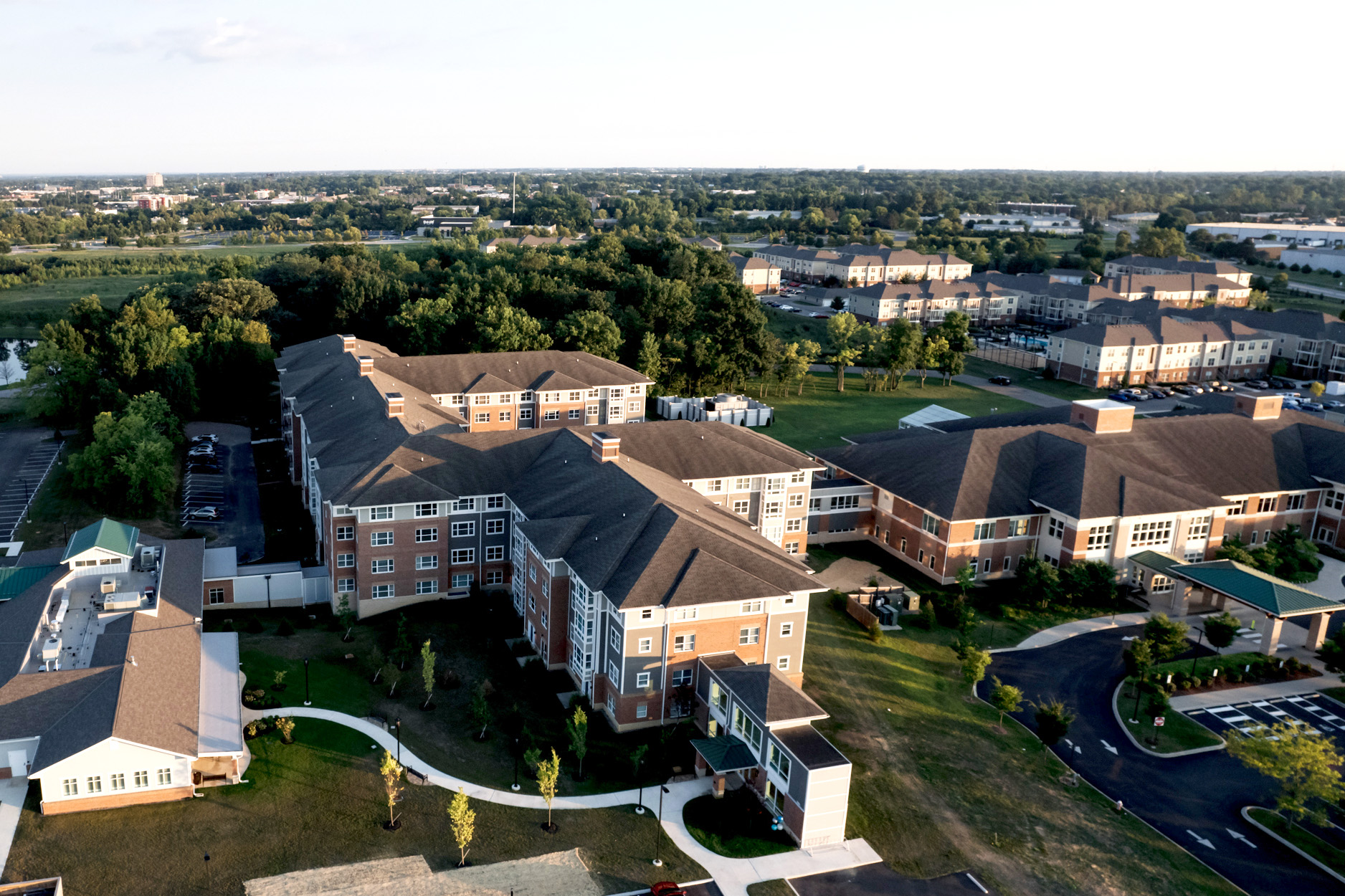Aerial view of a large residential complex with multiple connected brick buildings, surrounding green lawns, pathways, trees, and parking areas, set against a backdrop of a suburban landscape.