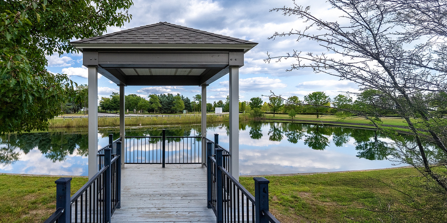 A wooden gazebo with a shingled roof overlooks a small pond surrounded by grass, trees, and cloudy blue sky reflected in the water. A railing lines the walkway leading to the gazebo over the pond.