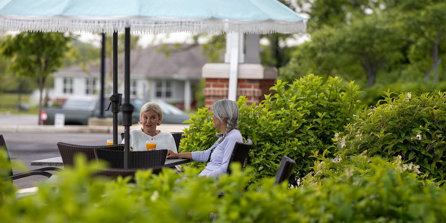 Two older women sit at an outdoor table under a large umbrella, talking and enjoying drinks, surrounded by lush green bushes with buildings and trees in the background.