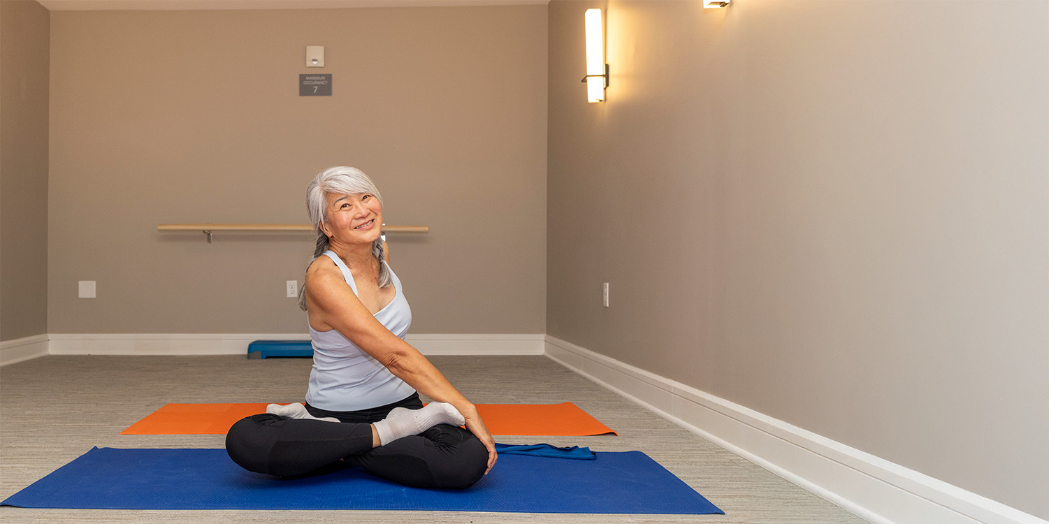 A woman with gray hair sits on a blue yoga mat in a cross-legged position, smiling and stretching her torso in a brightly lit room with beige walls and minimal decor.