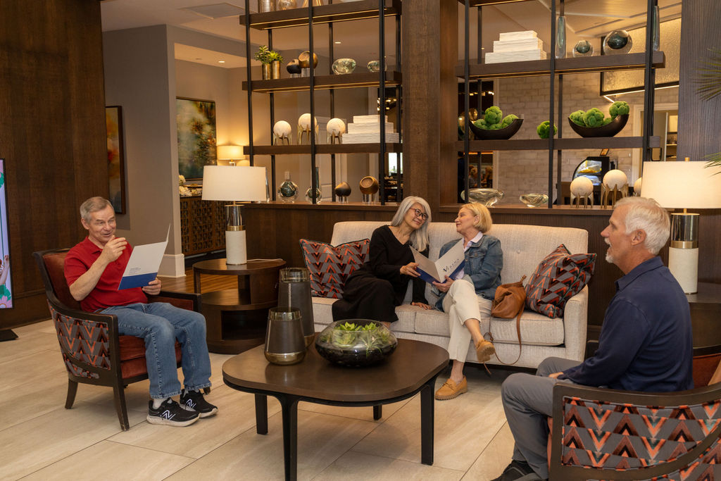 Four people relax and chat in a stylish lounge with modern decor and cozy seating. As one man reads a paper on Columbus home prices, the others, all 55+ homeowners, discuss plans to downsize and share smiles.