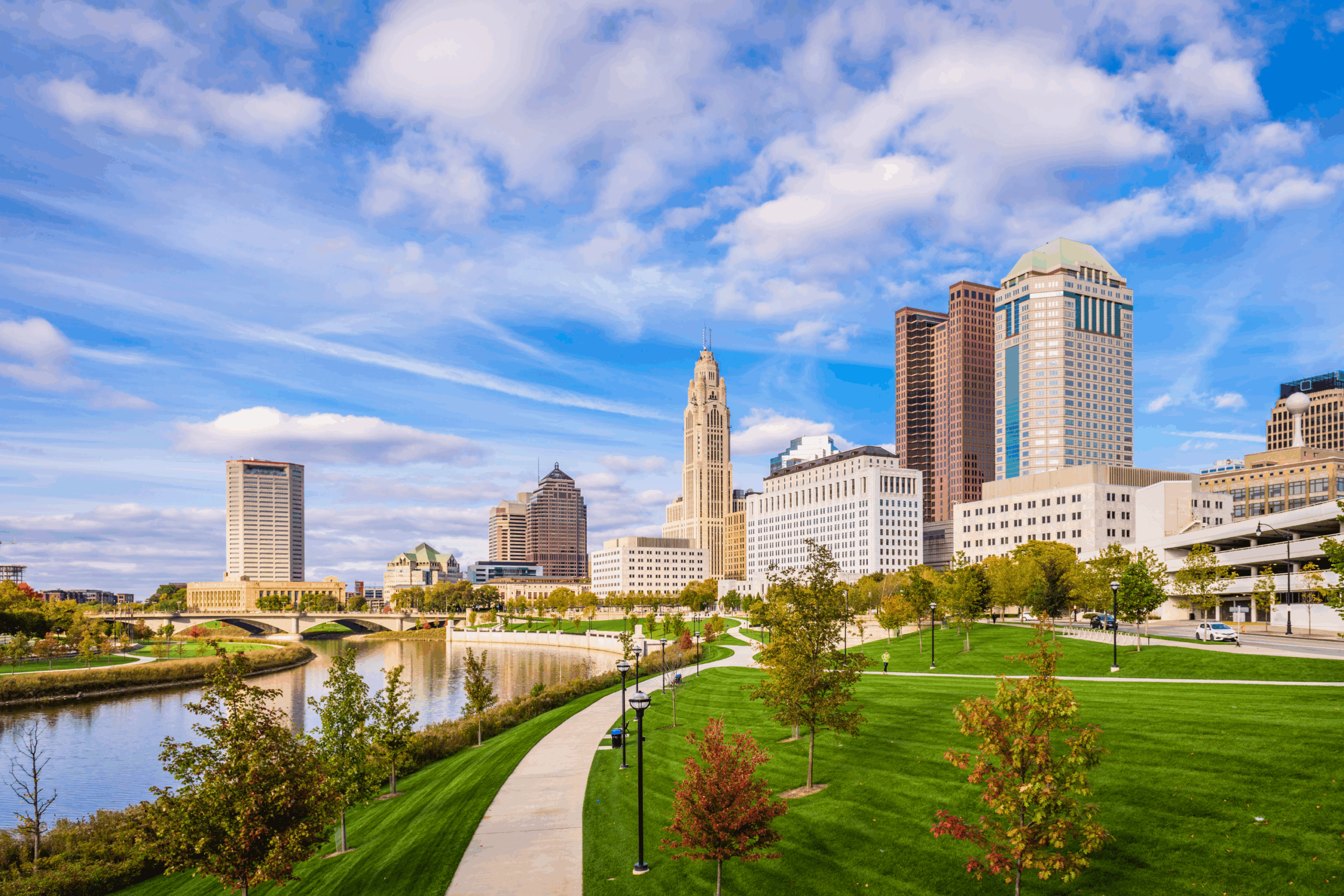 A city skyline with tall buildings and a river running alongside a green park with walking paths and trees under a partly cloudy sky, perfect for 55+ homeowners considering downsizing amid rising Columbus home prices.
