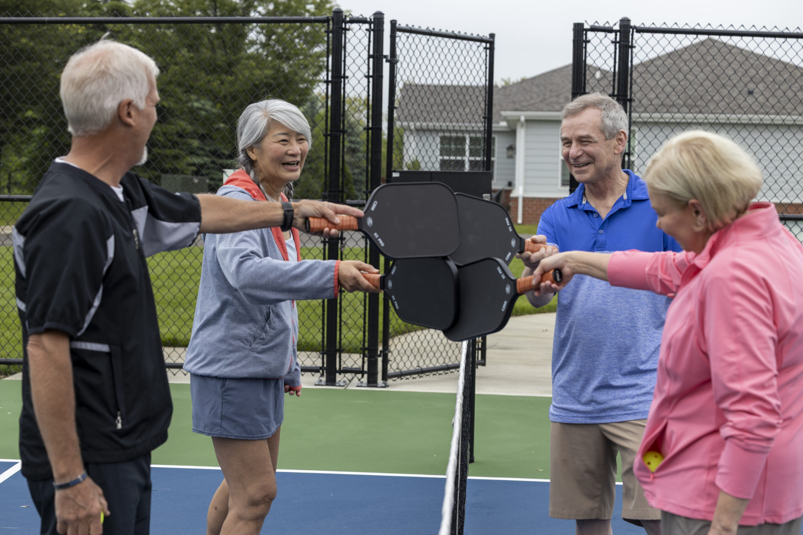 Four older adults stand on a pickleball court at The Cottages, smiling and holding their paddles together over the net in a gesture of camaraderie before a game. A fence and houses are visible in the background.