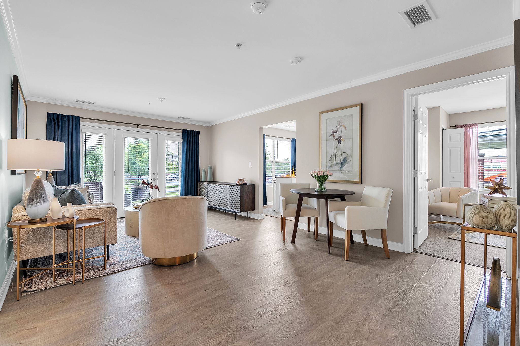 A bright, modern cottage living room with light wood floors, cream chairs, a small dining table, and large windows with blue curtains. An adjacent sitting area is visible through an open doorway.