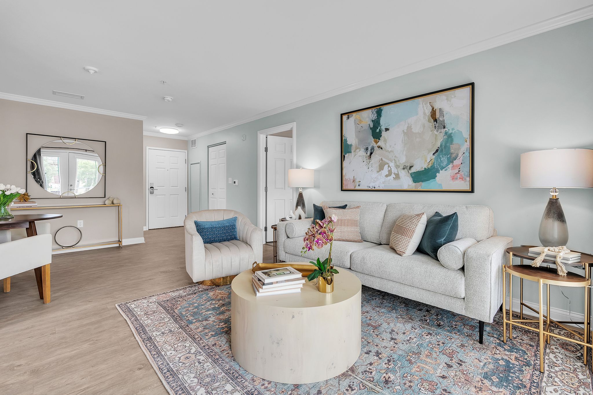 A modern living room with a light gray sofa, accent chair, round coffee table, and abstract wall art. The room has wood flooring, a patterned rug, and is decorated with plants and pillows. A console table and mirror sit against the wall.