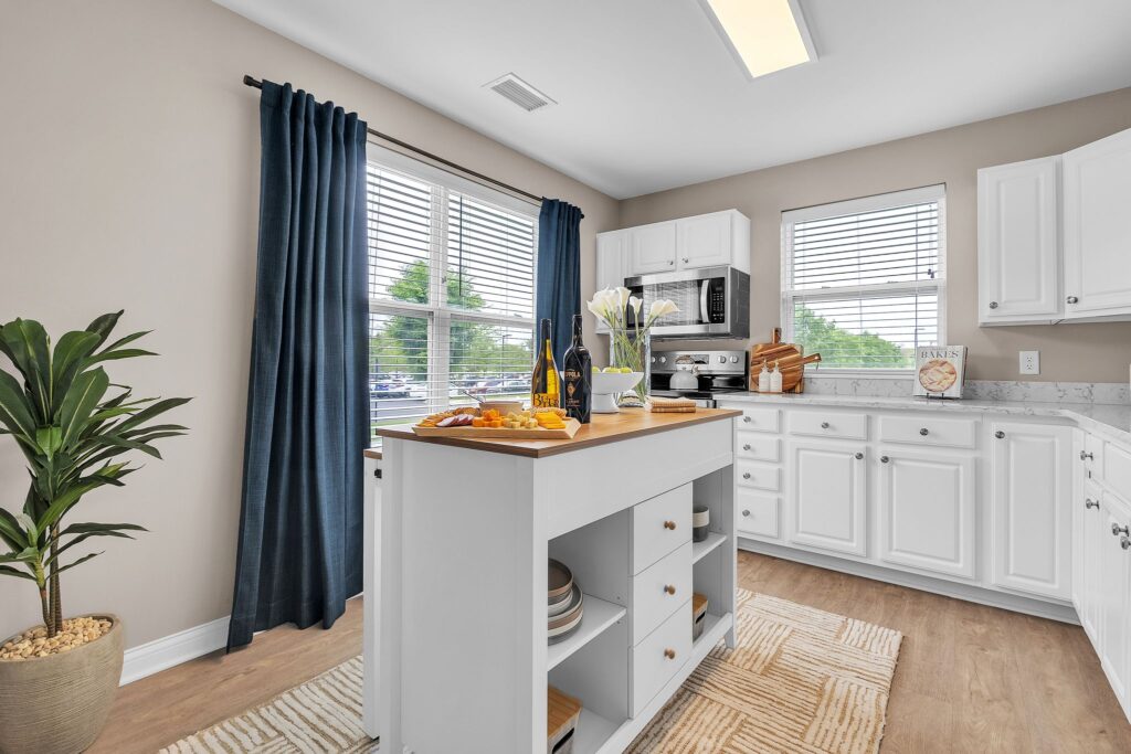 Bright kitchen in luxury apartments with white cabinets, a center island set with wine, glasses, and snacks, light wood flooring, two windows with dark curtains, a potted plant, and modern decor.
