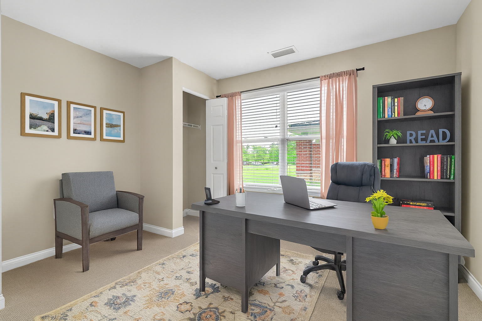 A home office in two-bedroom residences with a gray desk, laptop, black office chair, bookshelf with READ letters, armchair, framed art on the wall, and a window with pink curtains letting in natural light—live your way with more space.