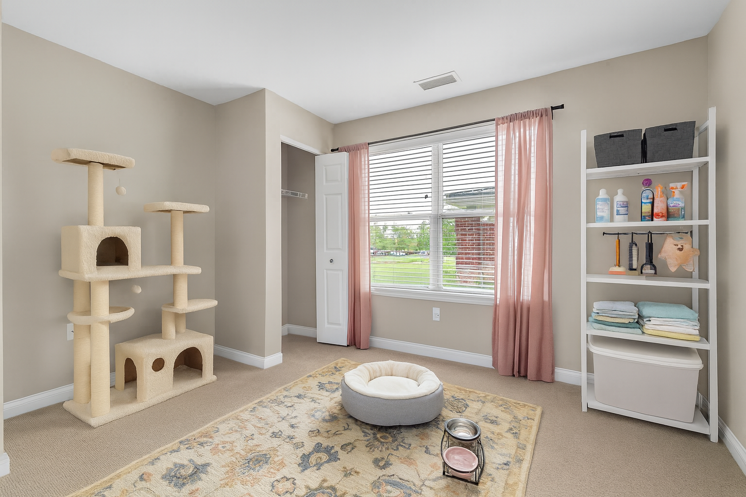 A tidy room with beige walls, a cat tree, a round pet bed, food and water bowls, and more space to live your way—perfect for pets. Includes a shelf with supplies and towels, an open closet, patterned rug, and a large window with pink curtains.